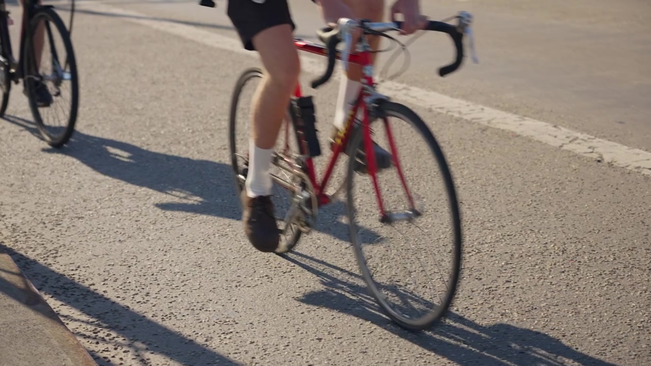 Nærbillede fra lav vinkel fanger en cyklist, der træder i pedalerne på en rød racercykel langs solrig asfalt, kaster skarpe skygger, mens det understreger fitness, bymobilitet og en aktiv livsstil
