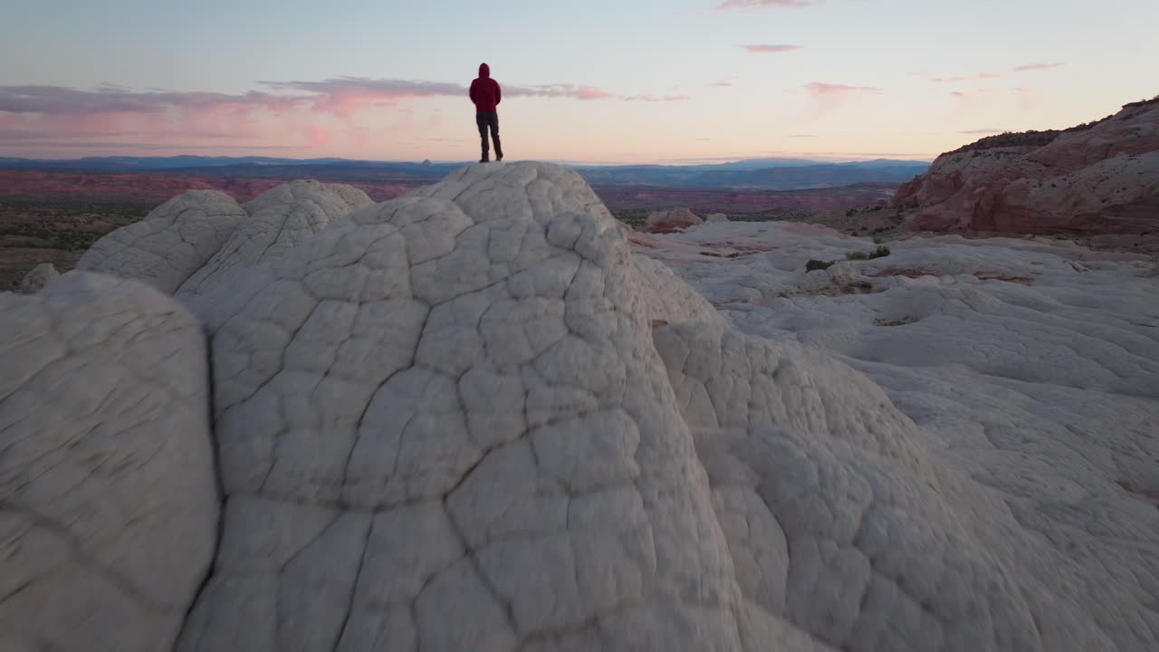 A drone flying forward and passing a male subject standing on top of the unique sandstone rock features of White Pocket Arizona surrounded by sandy desert and blue skies at blue hour sunrise