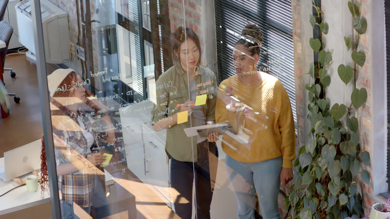Diverse female team collaborating on coding project, writing on glass wall in modern office