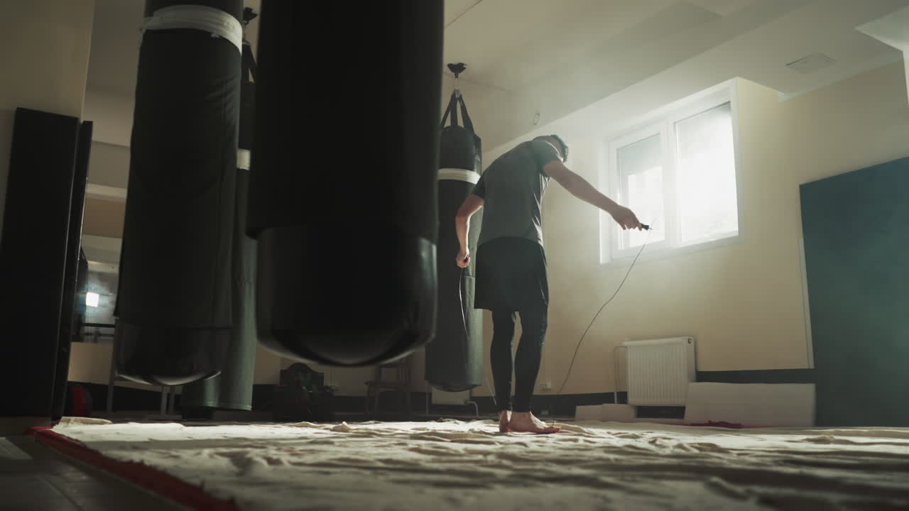 Fighter uses jump rope to train in gym. Young kick-boxer hops on skip rope in empty martial arts club slow motion. Athlete with exercise equipment