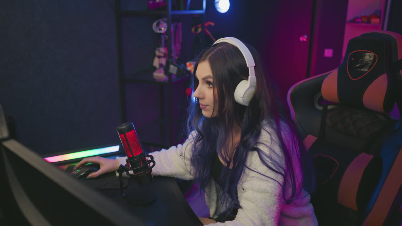 cheerful young woman wearing headphones adjusts forward with smile and fixes her hair after winning game, sitting at computer desk in cozy illuminated studio setup with microphone and gaming chair
