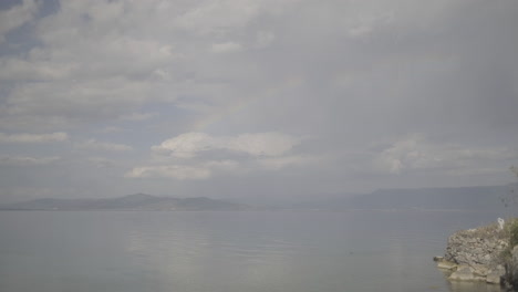 panning shot filmed from lin albania on a cloudy day revealing macedonia in the background with a beautiful rainbow forming in the clouds and blue water around log