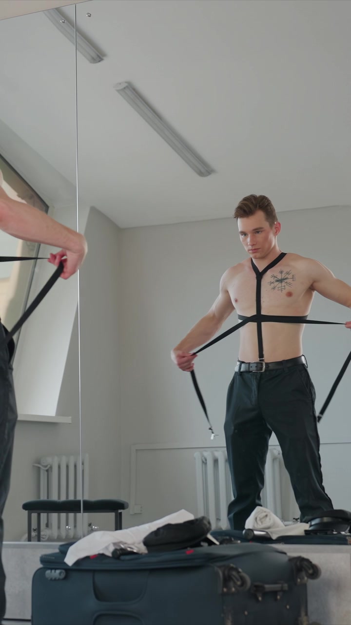 Shirtless young man with chest tattoo adjusts black suspenders. Room features large windows suitcases. Man secures straps around his torso while another man assists