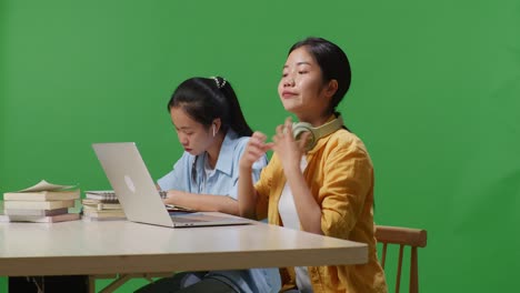 asian woman students wearing headphones and typing on a laptop while sitting with her friend studying on a table in the green screen background classroom