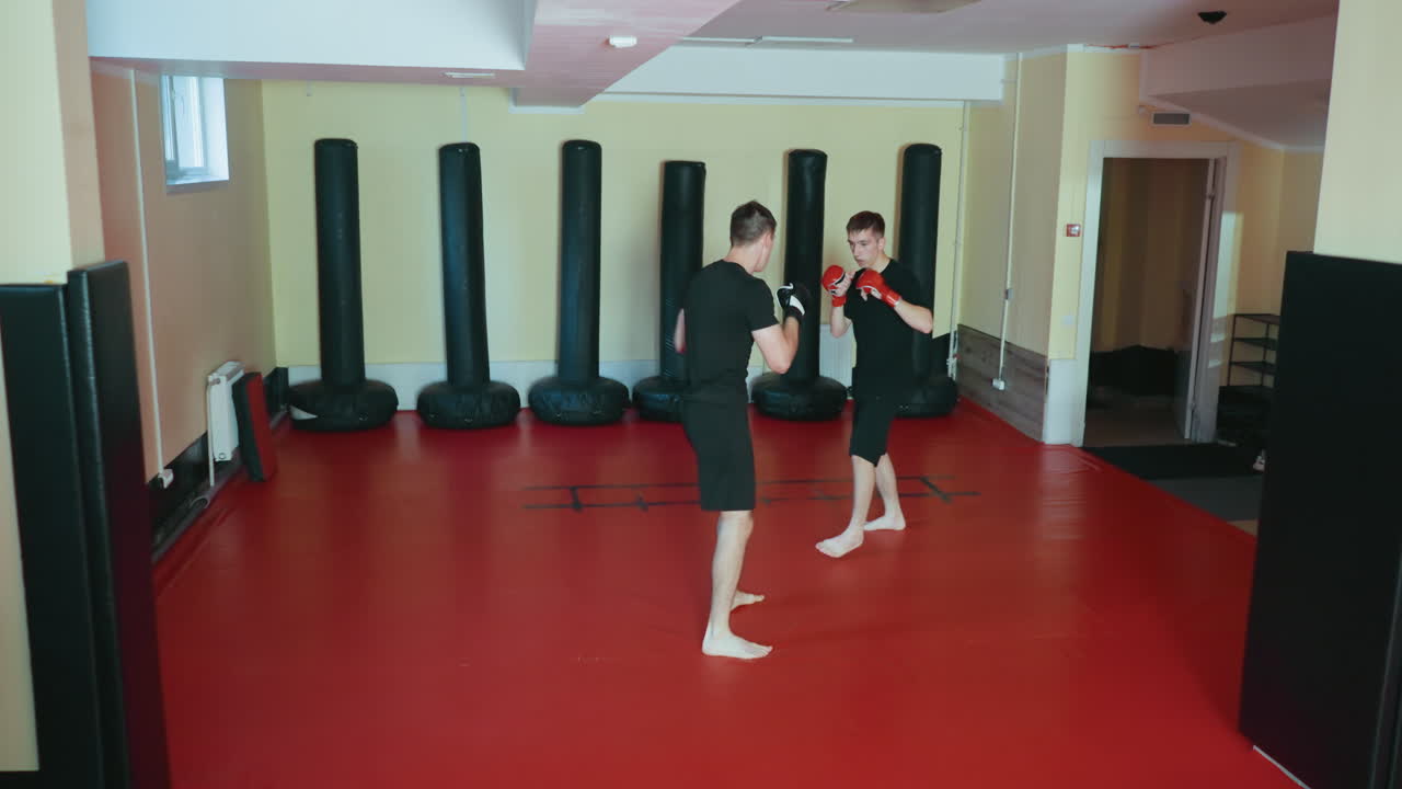 Athletes practicing combat sport sparring in martial arts gym on red mat floor, wearing black training outfits and gloves, performing boxing stance with punching technique near punching bags indoors