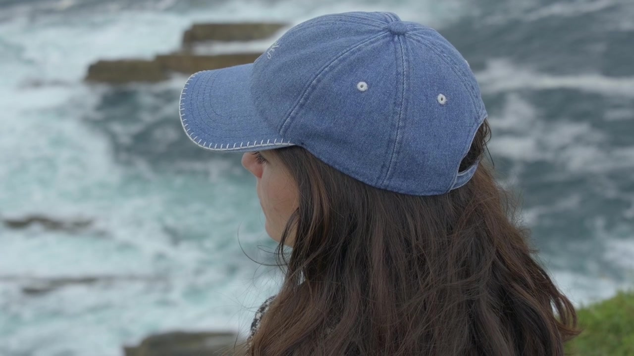 Back view of woman in denim cap watching ocean waves crash on rocks