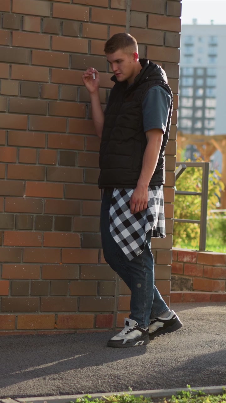 Primo piano di un giovane uomo appoggiato con disinvoltura a un muro di mattoni all'aperto, con una sigaretta in mano. L'uomo indossa un gilet nero sopra una camicia blu con una sovracamicia a quadri e jeans, mostrando una postura rilassata.