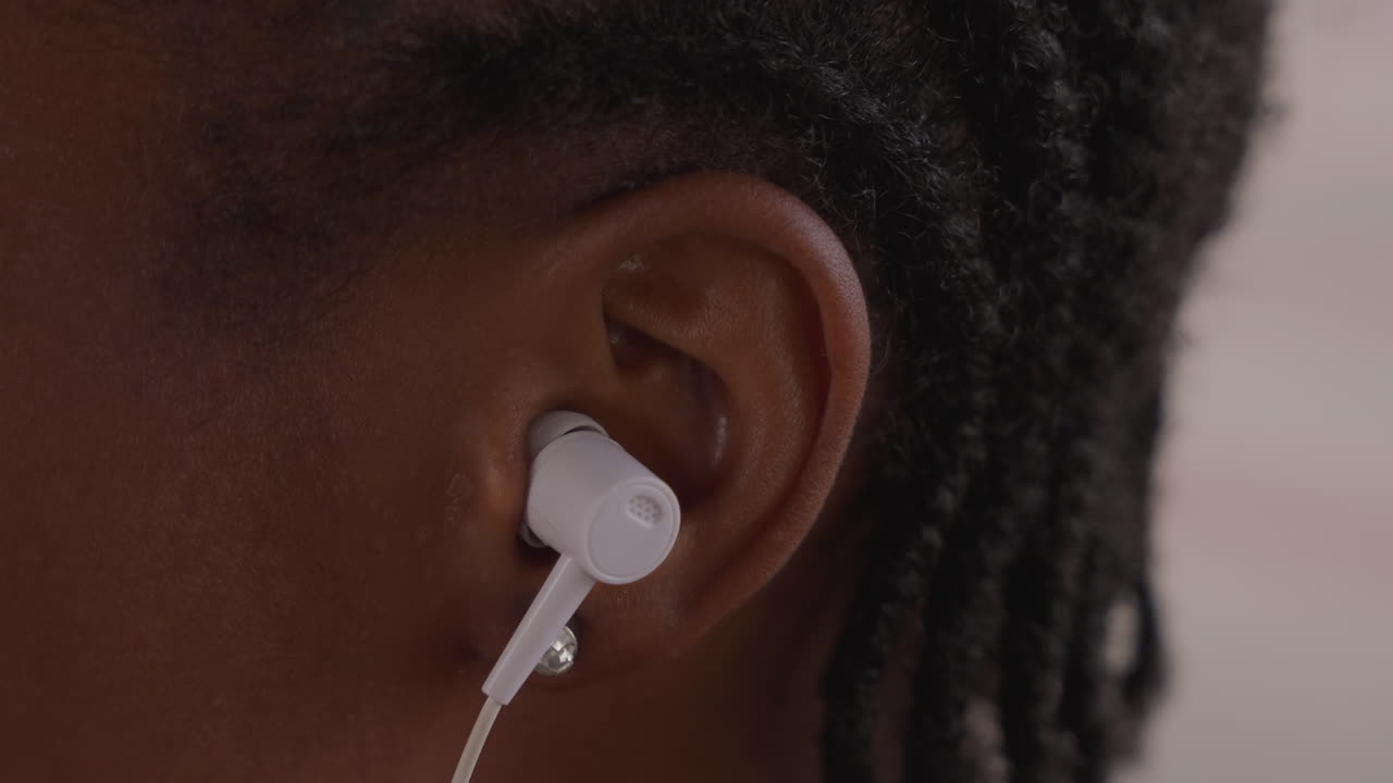 Gros plan sur un adolescent noir portant un écouteur, cheveux tressés et peau texturée, câble filaire visible, lumière naturelle douce, expression sereine et concentrée, suggérant une séance d'étude ou un auditeur en déplacement, moment personnel moderne