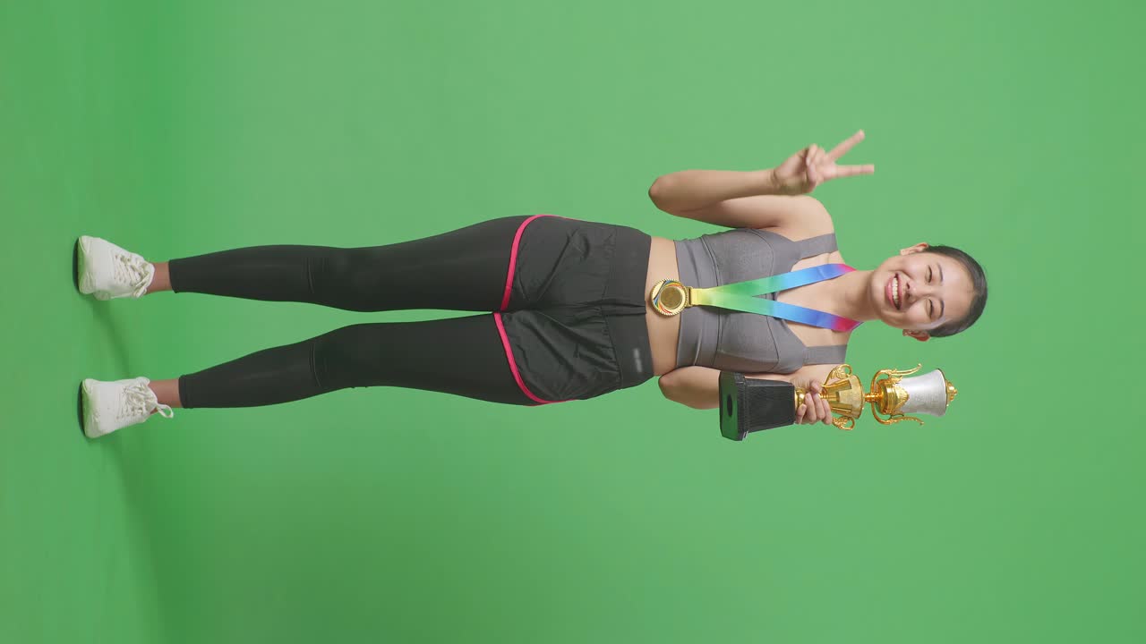 금메달과 트로피를 들고 있는 아시아 여성의 몸은 평화의 몸짓을 보여주고 스튜디오의 녹색 스크린 배경에서 첫 번째 우승자로 카메라에 미소 짓고 있습니다.