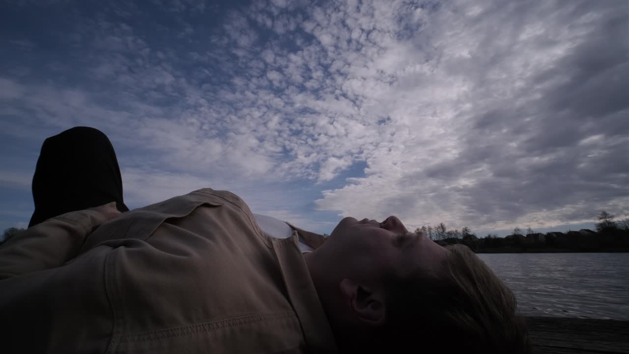 젊은이가 나무로 된 보행자 다리 위에 혼자 누워서 호수를 쳐다보고, 삶에 대해 생각하고, 자연에서 평화로운 분위기를 누리고, 신선한 공기를 즐기고, 뒤를 바라보고 있습니다.