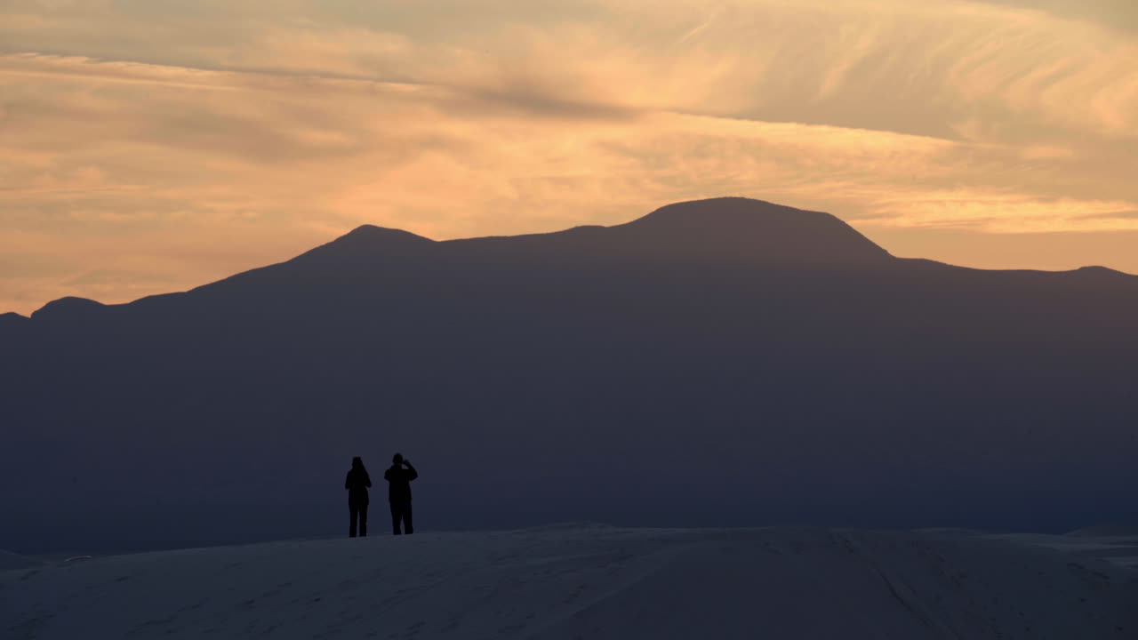 una pareja recortada en una duna de arena observa la puesta de sol sobre las montañas a lo lejos, 4k