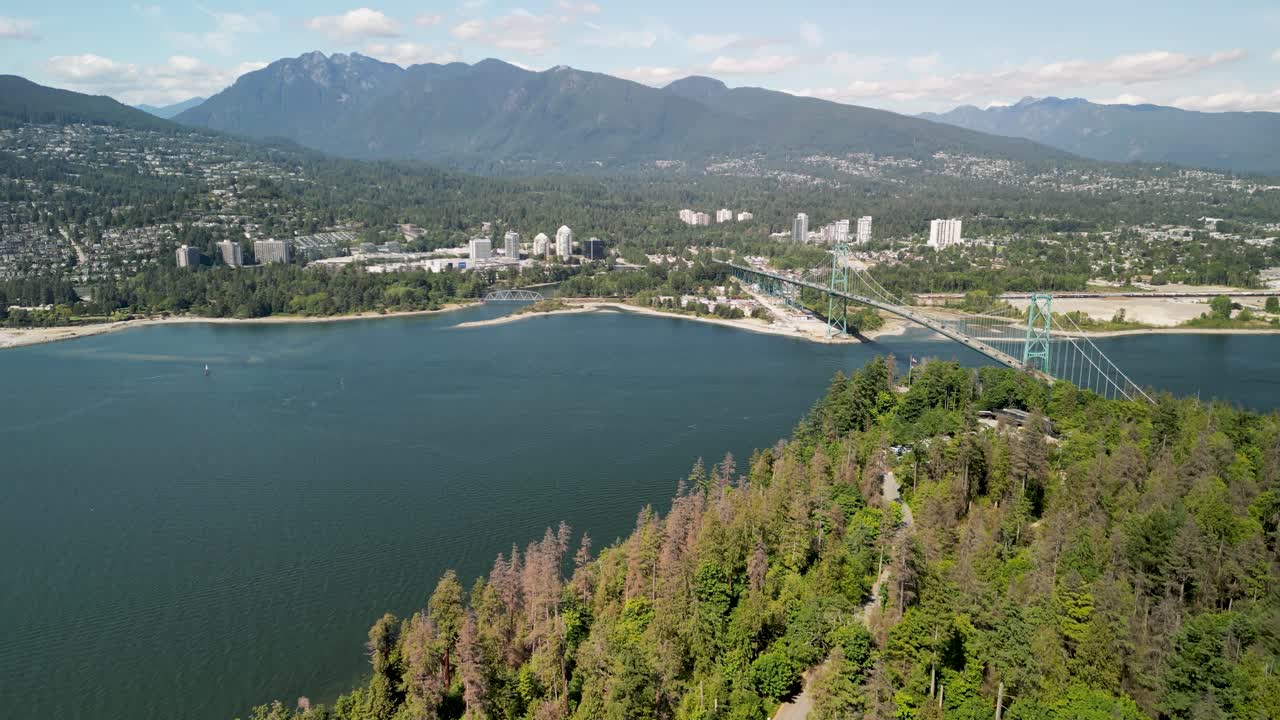 Riprese aeree con drone di Vancouver, con colline boscose in primo piano e lo skyline della città che si affaccia su acque costiere tranquille.