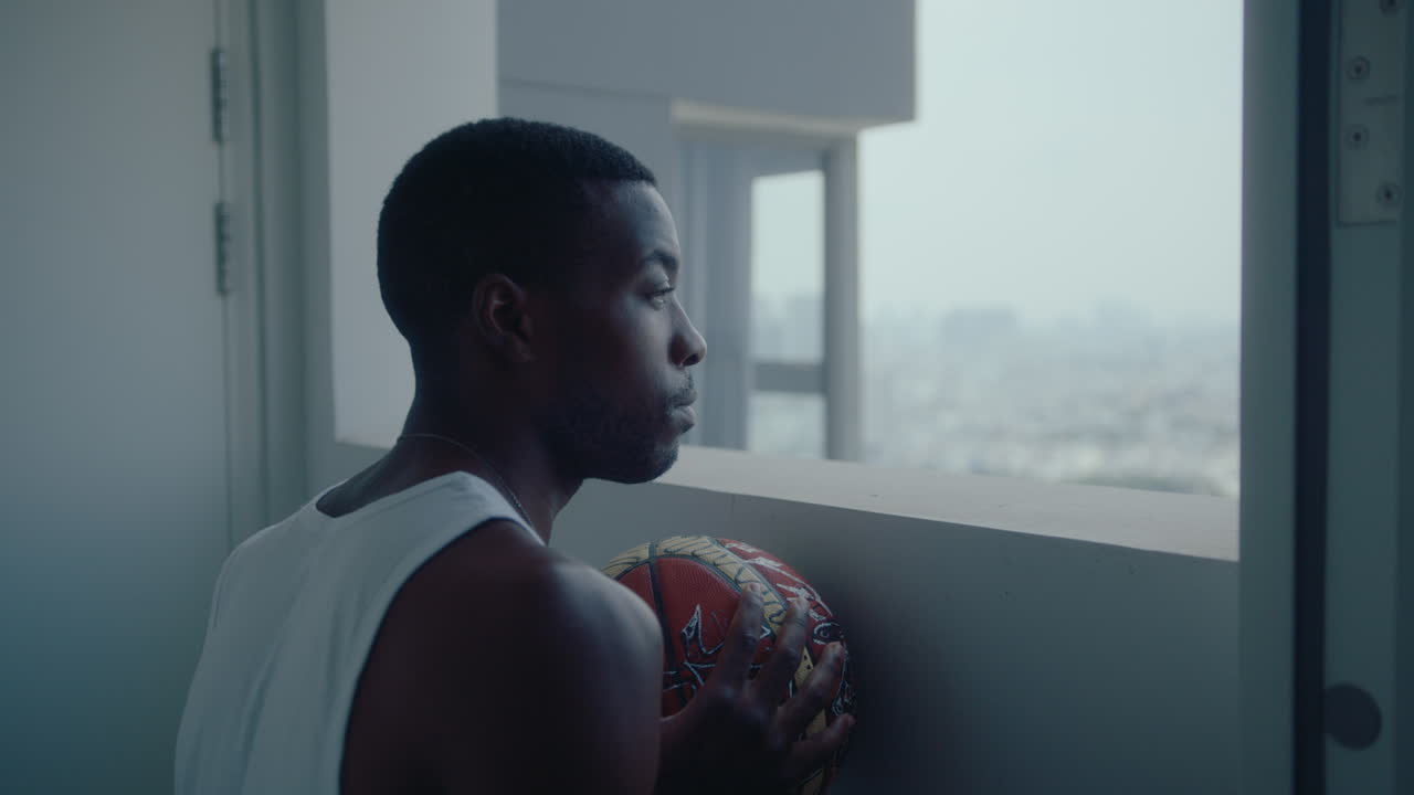 Homme noir avec un ballon de basket regardant le paysage urbain à travers la fenêtre du couloir