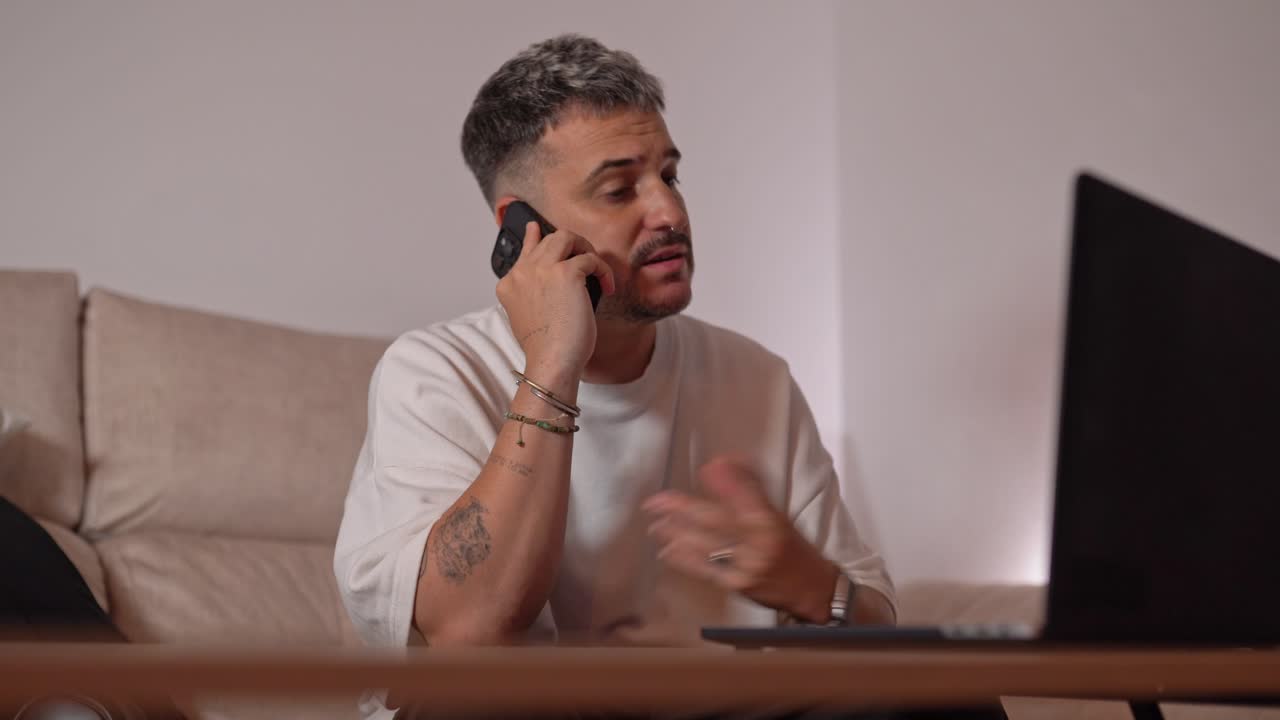 Angry man sitting on sofa shouting during intense phone call while gesturing towards laptop in home setting