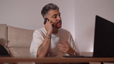 angry man sitting on sofa shouting during intense phone call while gesturing towards laptop in home setting