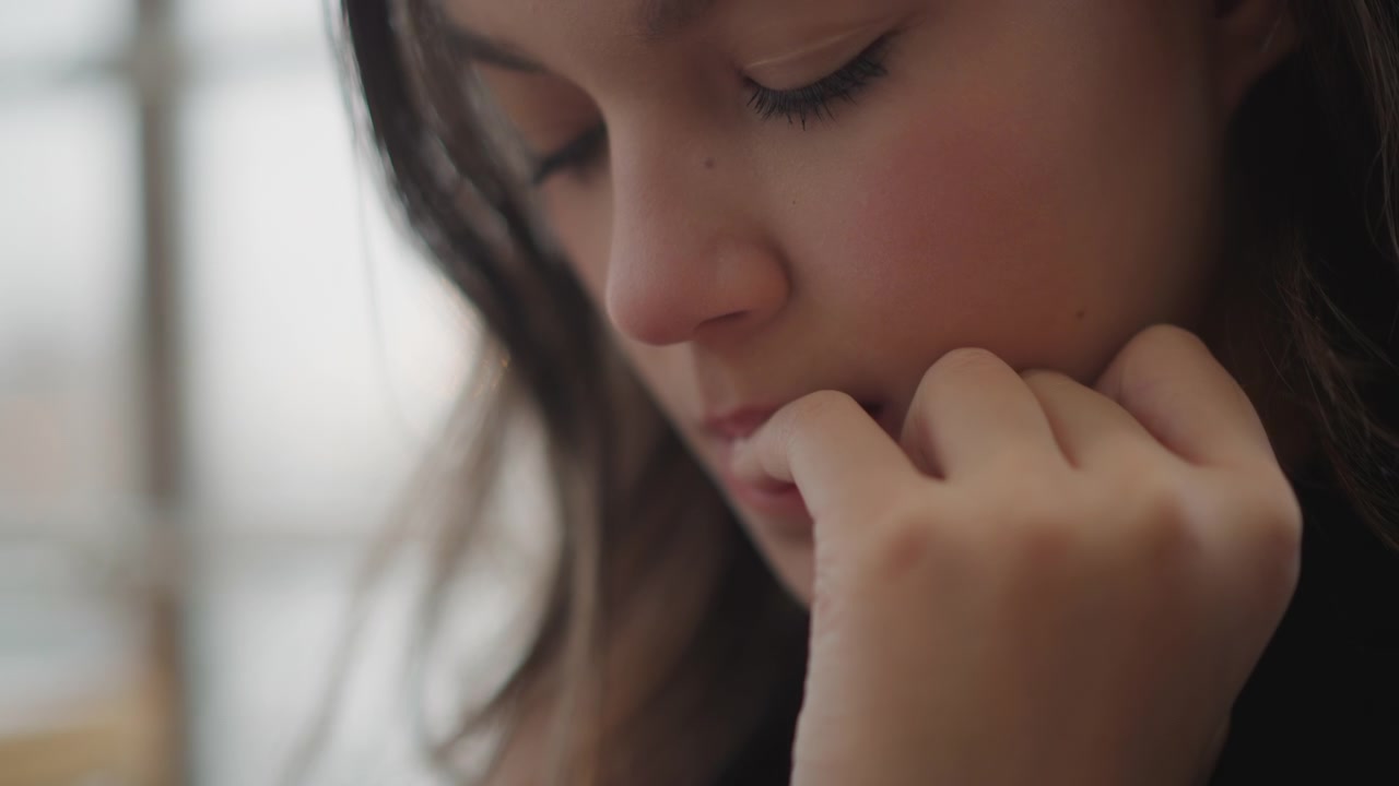 Tight indoor close-up of young woman bringing fingers to lips in small inward hand-to-mouth movement, with paused posture and soft light suggesting tension, hesitation, and quiet self-soothing