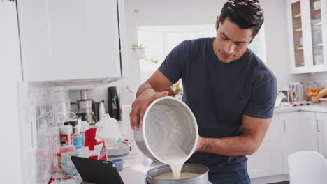 Millennial-man-pouring-cake-mix-into-a-form-following-a-recipe-on-a-tablet-computer,-close-up