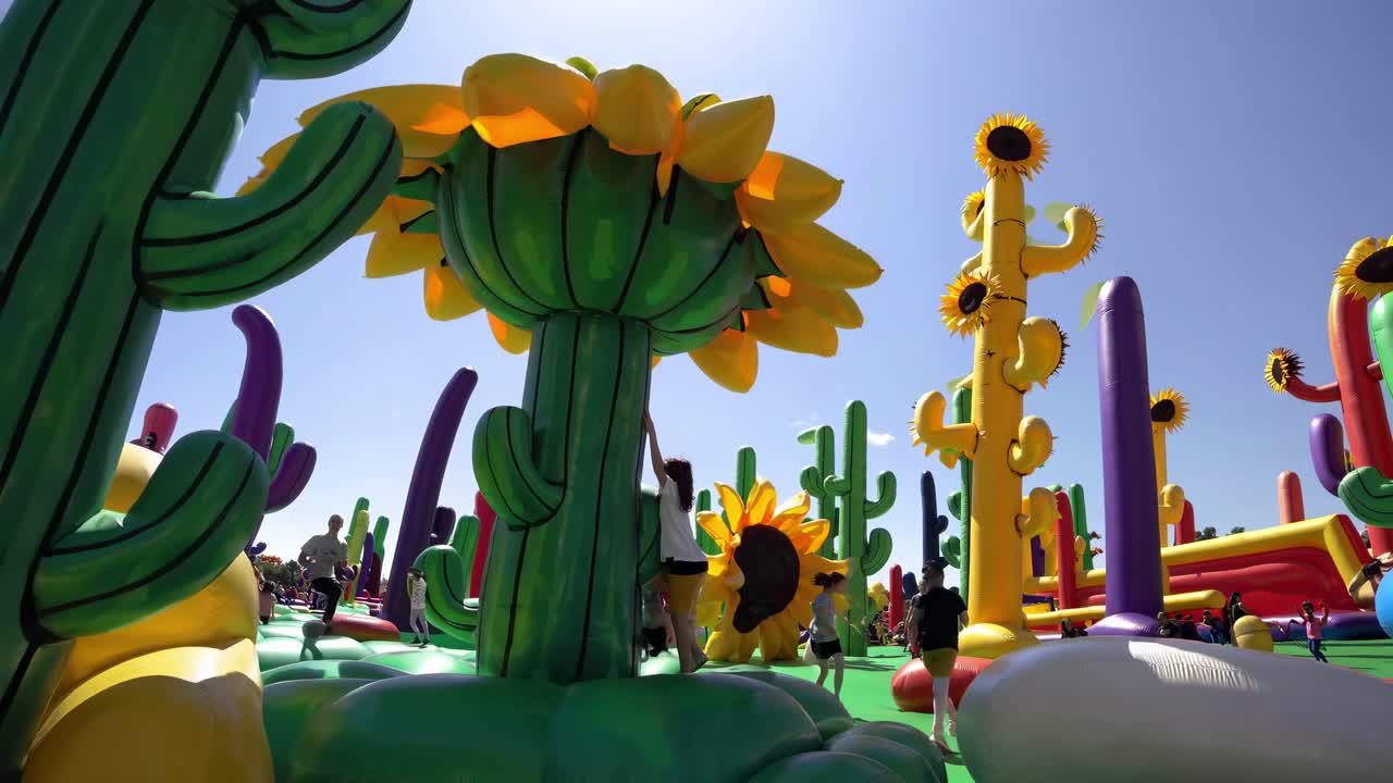 Kids enjoying jumping and climbing on colorful inflatable structures shaped like sunflowers and cacti in a vibrant outdoor playground, providing fun and entertainment