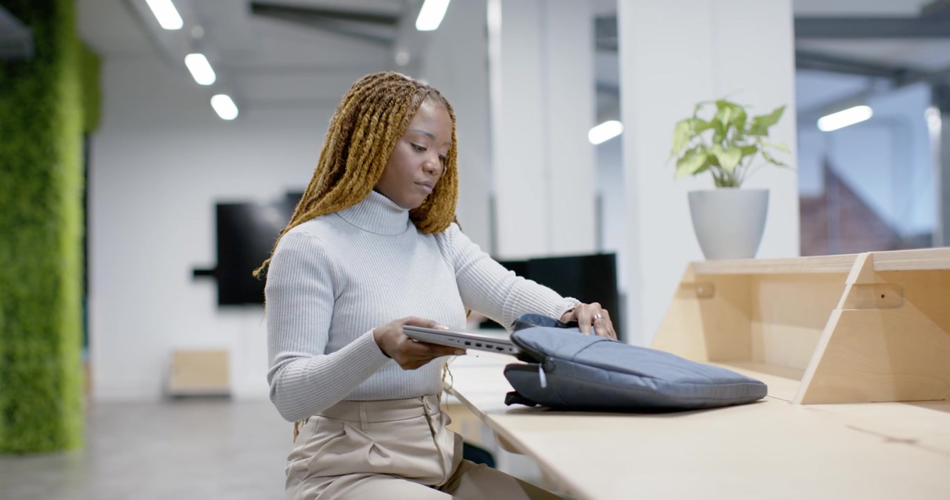 Femme d'une trentaine d'années en pull et pantalon, assise au comptoir, ouvrant un sac, sortant un ordinateur portable, déplaçant une tasse et se préparant.