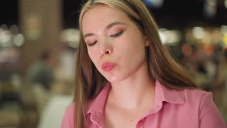 young woman seated in a mall, dressed in pink, eats a potato chip and then takes a drink, the background features a soft blur of glowing, diverse-colored lights with blur view of people