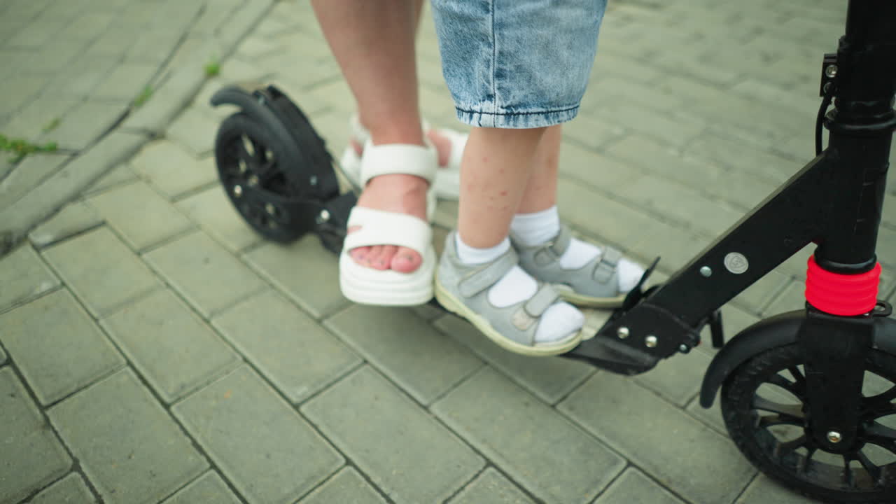 A woman hand is guiding a young boy's foot onto the scooter platform as he lifts his left leg