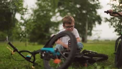 a young boy squats next to his bicycle on a grassy field, looking focused, with the partial view of someone else standing nearby
