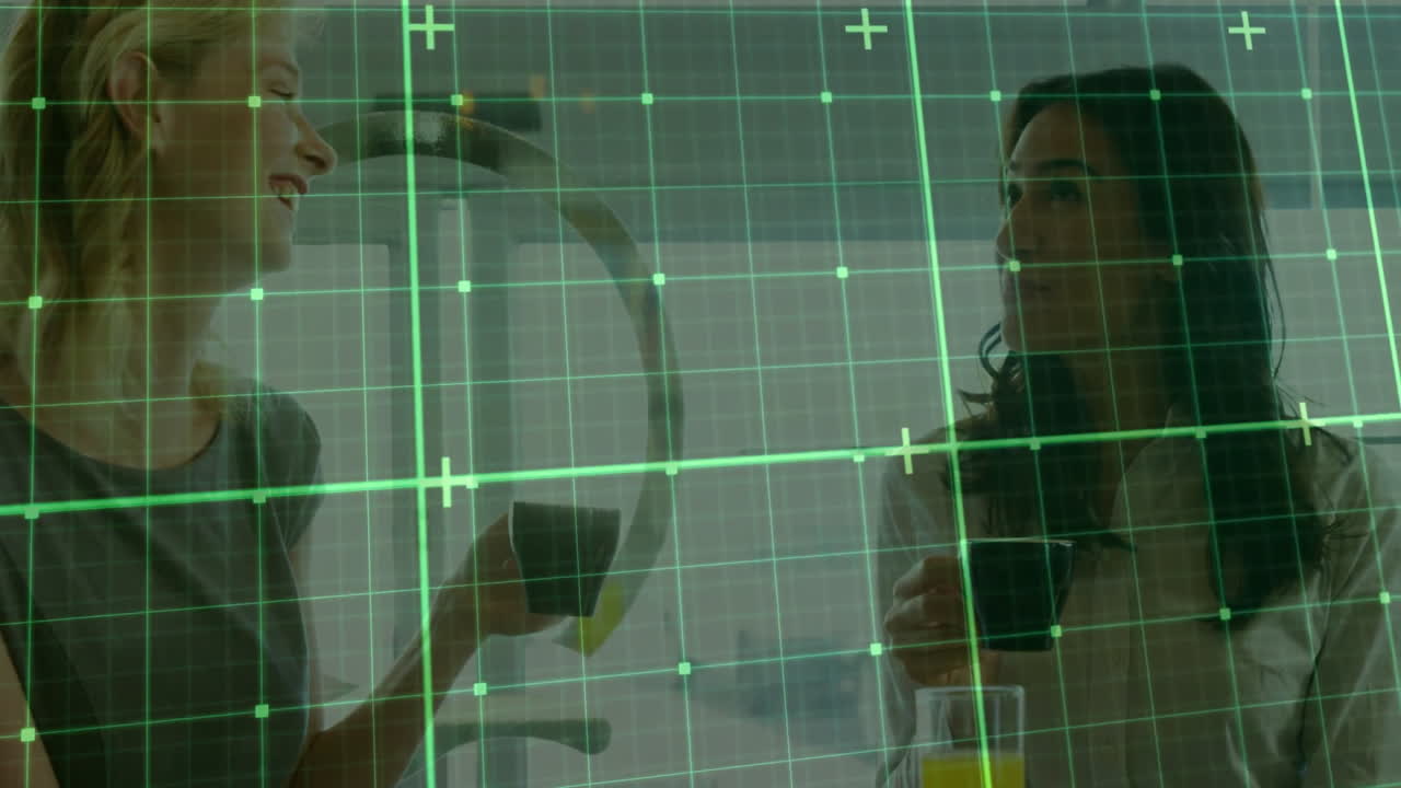 Two women sitting at table in tech lounge, holding coffee mugs and viewing digital grid overlay
