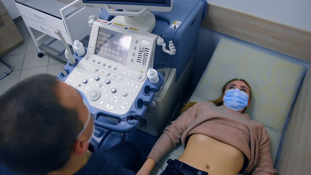 Blonde woman wearing mask lies on the doctor's couch. Male obstetrician uses ultrasonic device for patient's health examination. Top view.