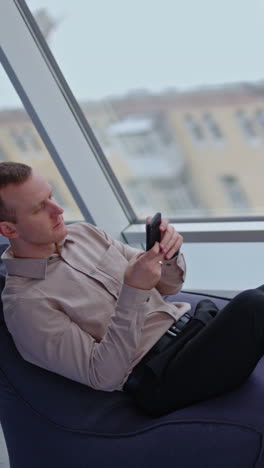 male worker resting in the bean bag chair. man searches information on the phone. panoramic window with city view at backdrop. vertical video