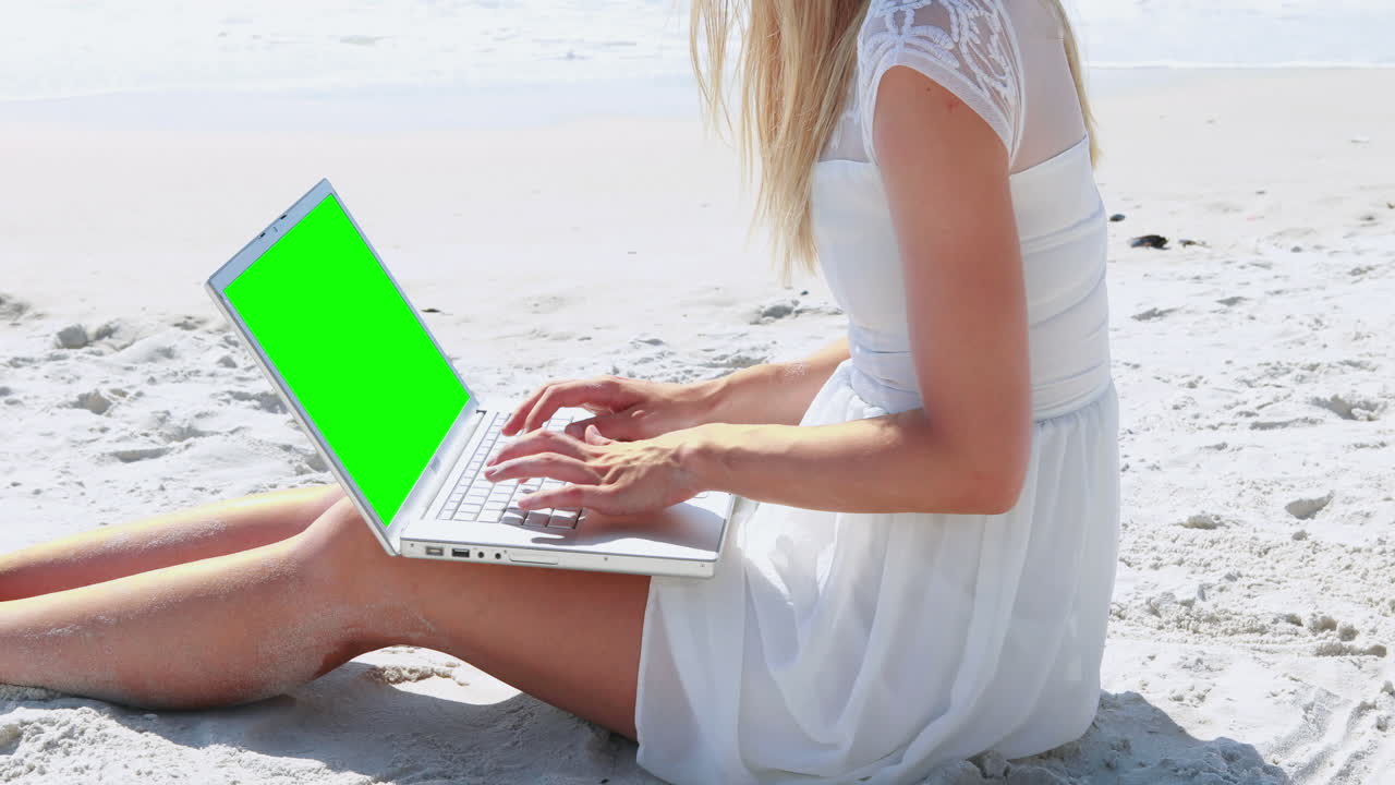 una mujer rubia usando una computadora portátil en la playa.