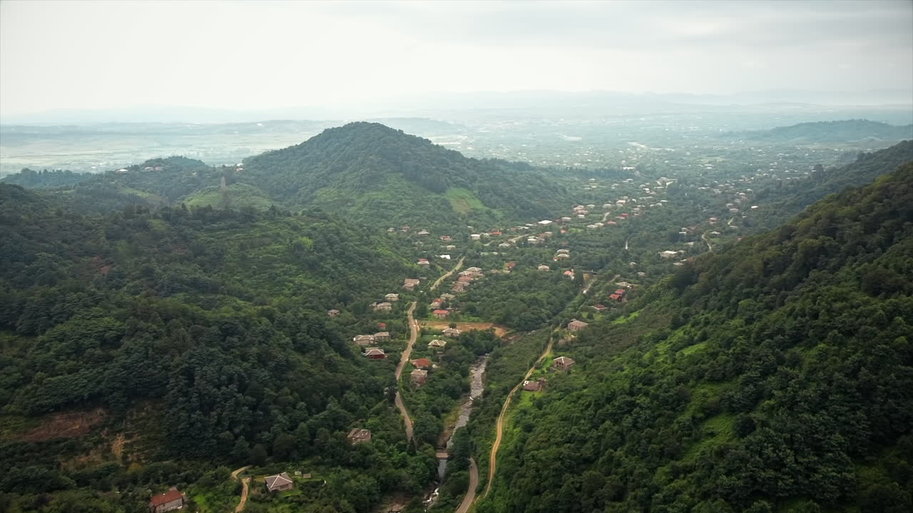 Dronebillede af naturen i Georgien. Dal med landsby, bakkede skråninger dækket af grønt, by i det fjerne og en skyet himmel.