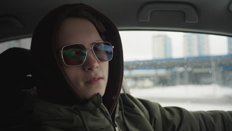 confident young man in hoodie and sunglasses sitting inside car with hand on wheel looking outside with slight smile as blurred snowy cityscape seen through car window in soft winter daylight