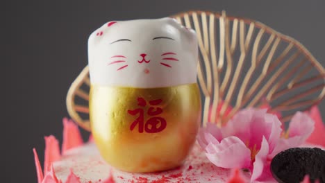 Figura-De-Maneki-neko-En-Un-Pastel-Con-Un-Ventilador-Y-Flores-De-Cerezo-En-Un-Fondo-De-Enfoque-Suave