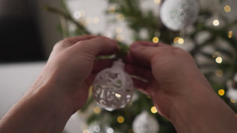 pov de manos colgando una delicada bola de navidad filigrana en un árbol con luces brillantes