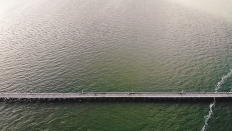 High,-tilting-camera-flyover-featuring-a-long,-wooden-pier-that-stretches-along-a-vast,-deep-blue-expanse-of-water-off-the-coast-to-a-beach-on-a-warm,-sunny-day