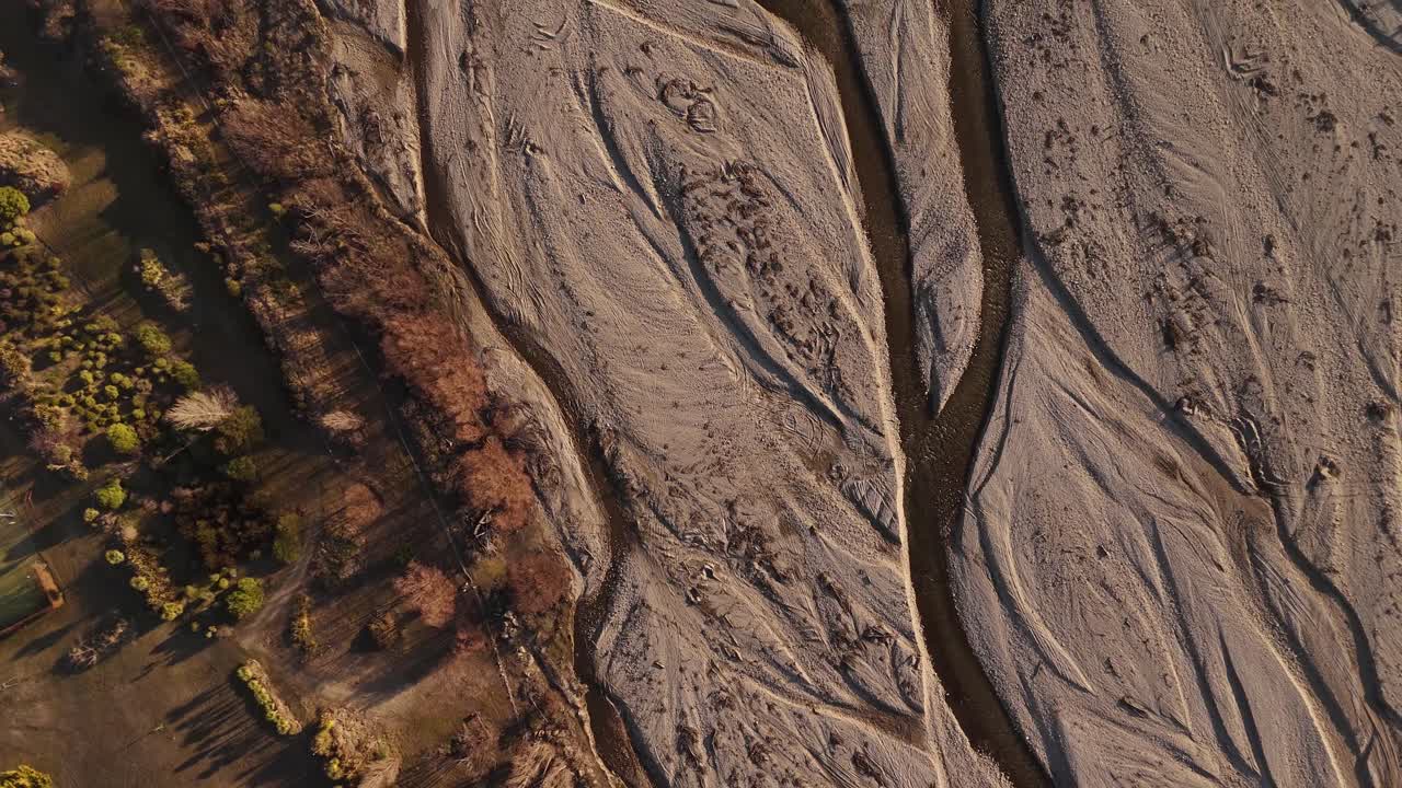 Cinematic top-down drone view of the Wanaka riverbed at sunset, highlighting the patterns of the dry river and flowing water in the Otago Region, New Zealand