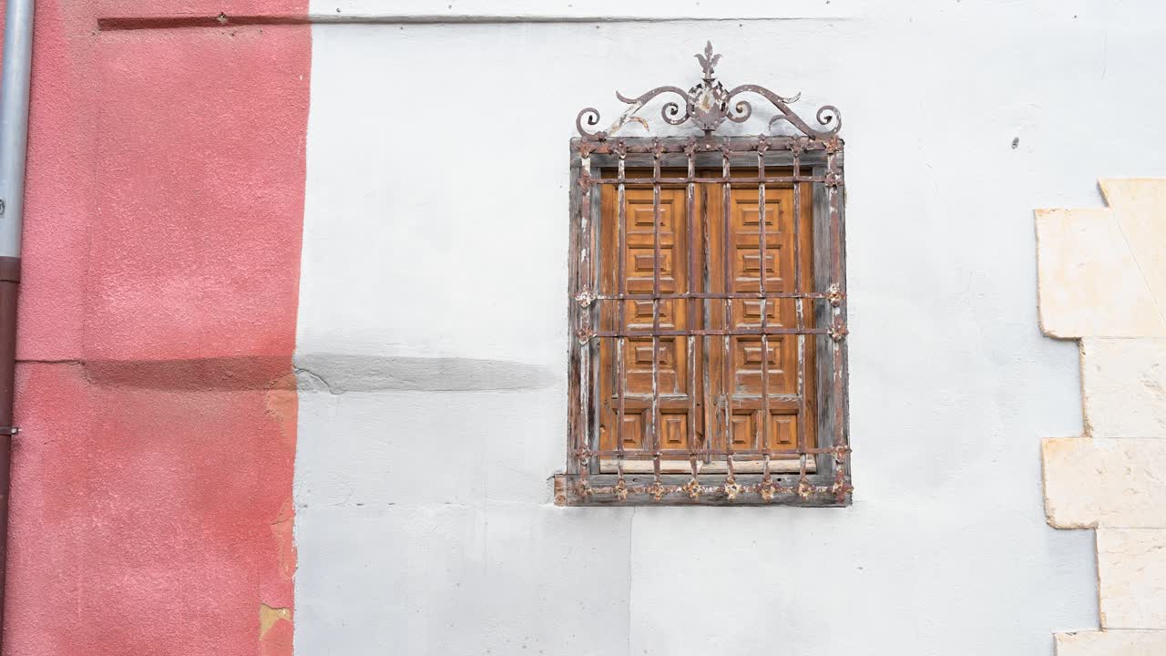 An architectural detail of a traditional house, featuring a window with wooden shutters protected by a classic, ornate wrought-iron grille in Cuenca, Spain.