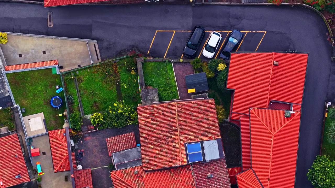 A direct overhead shot showing textured terracotta rooftops, small gardens, and parked cars in a village.