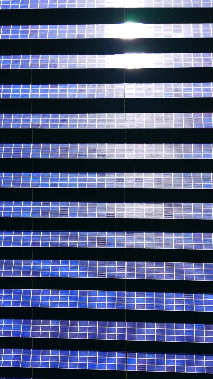 File ripetute di moduli solari visti dall'alto. Pannelli blu scuro riflettono la luce solare in brillanti fasce orizzontali attraverso l'impianto. Video verticale