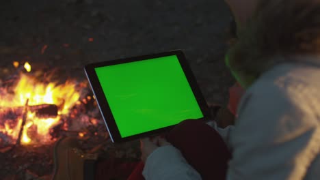girl is holding a tablet in landscape mode with green screen mock-up next to a campfire in the evening.