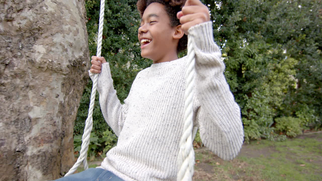 Happy african american boy swinging on tree swing in garden, in slow motion