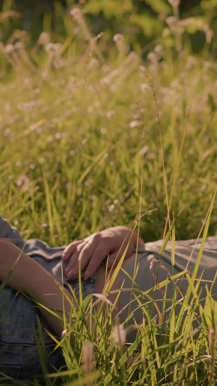 joven acostada en un campo de hierba, relajada con una mano apoyada en su pierna y otra en su estómago, rodeada de hierba alta y bañada en la cálida luz del sol