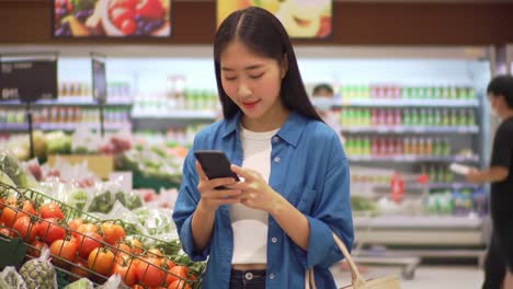 A young woman shopping in a grocery store, focused on her smartphone while surrounded by fresh produce and vibrant grocery items, merging modern technology with daily errands.