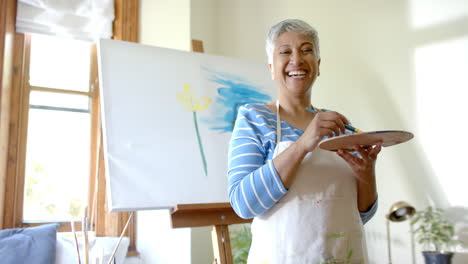 retrato de una feliz mujer biracial mezclando colores en la paleta en casa, en cámara lenta