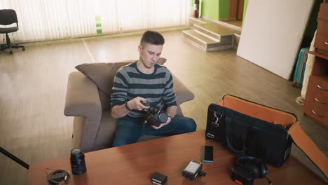 man sitting on sofa reaching for camera equipment bag on table. various gear visible such as lens, headphones, and smartphone. studio-like setup with a clean workspace and green walls