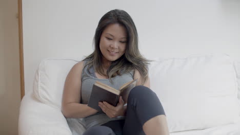 front view of cute woman sitting on sofa and reading book