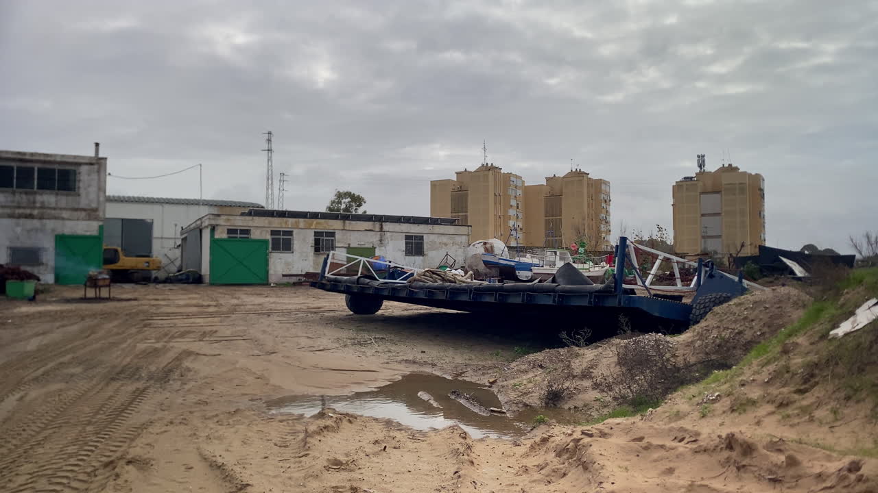 Sanlucar de Barrameda's coastal area featuring industrial buildings, an excavator, abandoned equipment, and apartment blocks under a cloudy sky