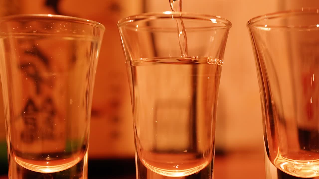 Primo piano di sake trasparente che viene versato in un bicchiere sotto una calda illuminazione ambientale da ristorante.