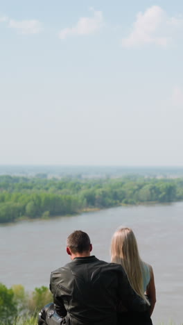 lovely couple of blonde lady and motorcyclist man sits resting on motorcycle on green hill near tranquil river on spring day backside view slow motion