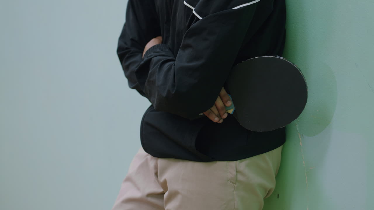 Close up partial view of tennis player in black jacket and beige pants leaning against wall with arms folded holding racket, relaxed stance highlighting patience